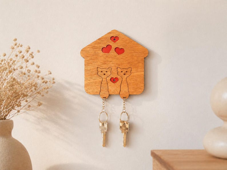 May include: Wooden key holder shaped like a house, featuring two cat silhouettes and red heart cutouts. Two sets of keys hang from the cat figures. The key holder is mounted on a light-colored wall, with a vase of dried flowers visible.