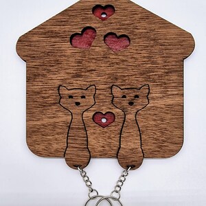 May include: Wooden key holder shaped like a house, featuring two cat silhouettes and three heart cutouts. The key holder is made of brown wood with red accents and includes two silver key rings.