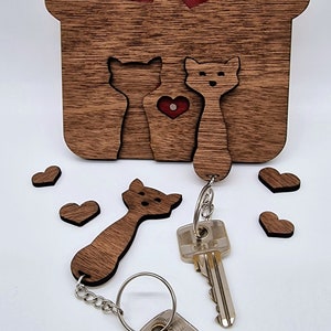 May include: Wooden key holder shaped like a house, featuring cat silhouettes and heart cutouts. Includes two cat-shaped keychains and several small heart-shaped accents. The key holder is approximately 6 inches tall.