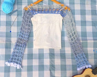 Op de afbeelding: Een witte top met een blauw en wit gehaakte overlay, weergegeven op een houten hanger. De gehaakte mouwen hebben een gradiënteffect, dat overgaat van lichtblauw naar een donkerdere tint. De manchetten zijn afgewerkt met wit kant. Een bolletje blauw garen en een ukelele zijn ook zichtbaar.