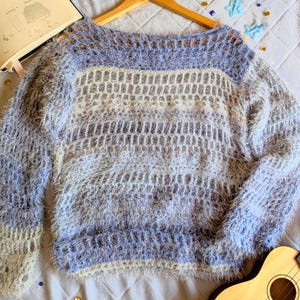 Op de afbeelding: Een lichtblauwe en witte fuzzy trui met een uniek open geweven ontwerp. De trui wordt getoond op een houten hanger. Een ukelele en een notitieboek zijn ook zichtbaar in de afbeelding.