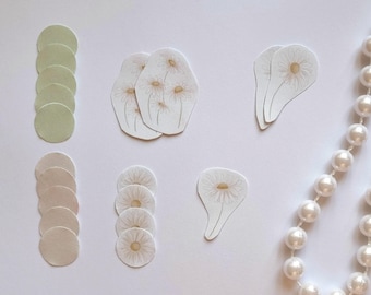 Op de afbeelding: Assortiment van witte en crèmekleurige papieren cirkels en bloemvormige stickers. De stickers hebben een delicaat bloemenmotief met een enkele bloem op elke sticker. De cirkels zijn gestapeld in twee stapels, een met een lichtgroene kleur en de andere met een crèmekleur.