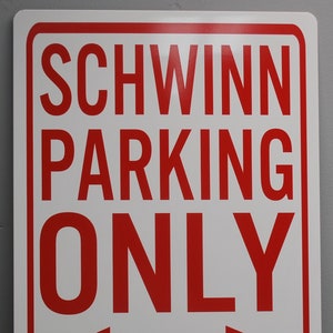 May include: Rectangular white sign with a red border and red text. The sign reads "SCHWINN PARKING ONLY" with an arrow pointing both directions. The bottom of the sign reads "ALL OTHERS WILL BE TOWED."