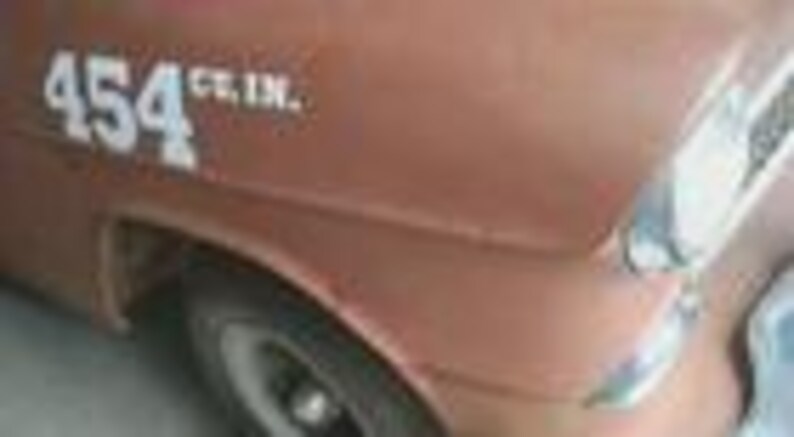 May include: Close-up of a vintage brown car's front quarter panel. White text reads "454 cu. in." on the side. The car has a chrome headlight and a black tire with a white rim. The paint shows signs of age.