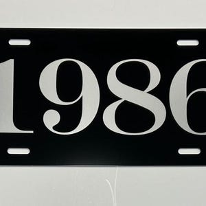 May include: Black license plate with the number "1986" in a bold, white font. The plate has four mounting holes and a glossy finish. The design is simple and modern.
