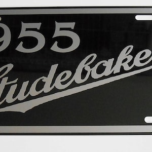 Könnte beinhalten: Schwarzes Nummernschild mit dem Text "1955 Studebaker" in silberner Schrift. Das Schild hat einen silbernen Rand und vier Befestigungslöcher. Klassisches Autozubehör.
