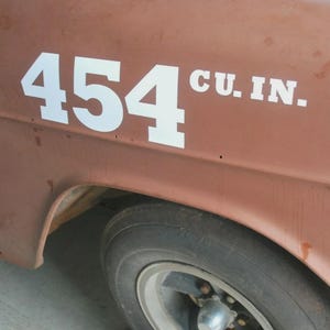 May include: Close-up of a classic car's side, featuring the white text "454 CU. IN." on a weathered, reddish-brown surface. A portion of the black tire and silver rim is visible, suggesting a vintage vehicle.