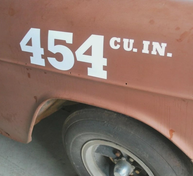 May include: Close-up of a classic car's side, featuring the white text "454 CU. IN." on a weathered, reddish-brown surface. A portion of the black tire and silver rim is visible, suggesting a vintage vehicle.