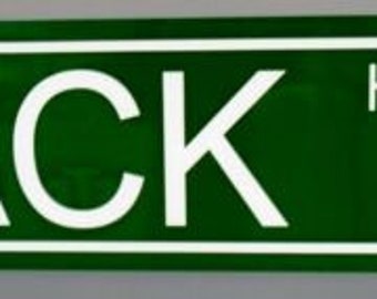 Op de afbeelding: Groen rechthoekig bord met witte rand en tekst. Het woord "MACK" staat prominent in grote witte hoofdletters aan de linkerkant, met "HIGHWAY" in kleinere witte letters aan de rechterkant.