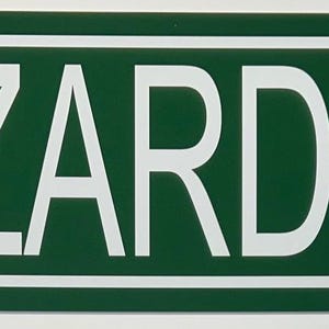 Puede incluir: Un letrero de calle verde y blanco con la palabra "HAZZARD" en letras mayúsculas blancas. El letrero también indica "3 MI." con una flecha blanca hacia arriba, que indica una dirección.