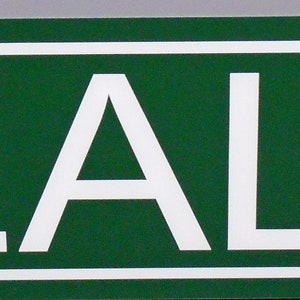 以下が含まれることがあります： 白い縁取りと白い文字で「REAL ST.」と書かれた緑色の長方形の道路標識。この標識は、大きな大文字で書かれた「REAL」という単語と、右上に配置された「ST.」という略語を備えた、すっきりとしたフラットなデザインが特徴です。