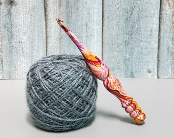 May include: A crochet hook with a marbled, swirling pattern of orange, pink, red, and white colors, featuring a contoured, ergonomic handle with a series of rounded ridges, positioned next to a round ball of textured, medium-gray yarn.
