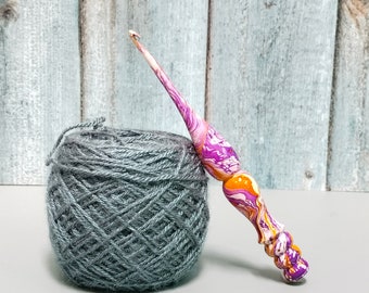 May include: A crochet hook with a marbled handle featuring swirls of purple, orange, and white, positioned next to a round ball of grey yarn. The hook has a tapered metal tip and a contoured, multi-textured handle with a glossy finish.