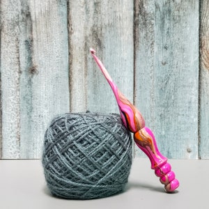 May include: A crochet hook with a smooth, ergonomic handle featuring a marbled pattern of bright pink, orange, black, and white swirls, positioned next to a round ball of dark grey yarn with a visible textured fiber construction.