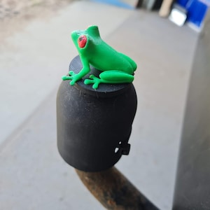 May include: A green frog figurine with red eyes sits on top of a black bottle stopper.