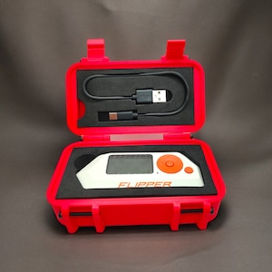 May include: A red hard case with a white and orange device inside. The device has a screen and a button. The case is open and the device is visible. The text "FLIPPER" is printed on the device.
