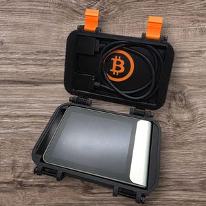 May include: Open black case with orange accents, holding a black cable and a device with a screen. The case features a Bitcoin symbol in orange. The device has a white edge. The case is open on a wooden surface.