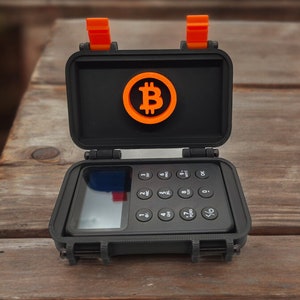 Può includere: Una piccola custodia nera con chiusure arancioni è aperta per rivelare un piccolo dispositivo nero con un display digitale e una tastiera. Il dispositivo ha un simbolo Bitcoin nella parte superiore.