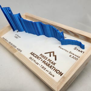May include: A blue 3D topographical map of Pikes Peak, part of the Ascent + Marathon, set in a wooden frame. The map shows the elevation profile and includes text indicating the start at Manitou Springs, 6,300 feet, and the 26.2-mile course.