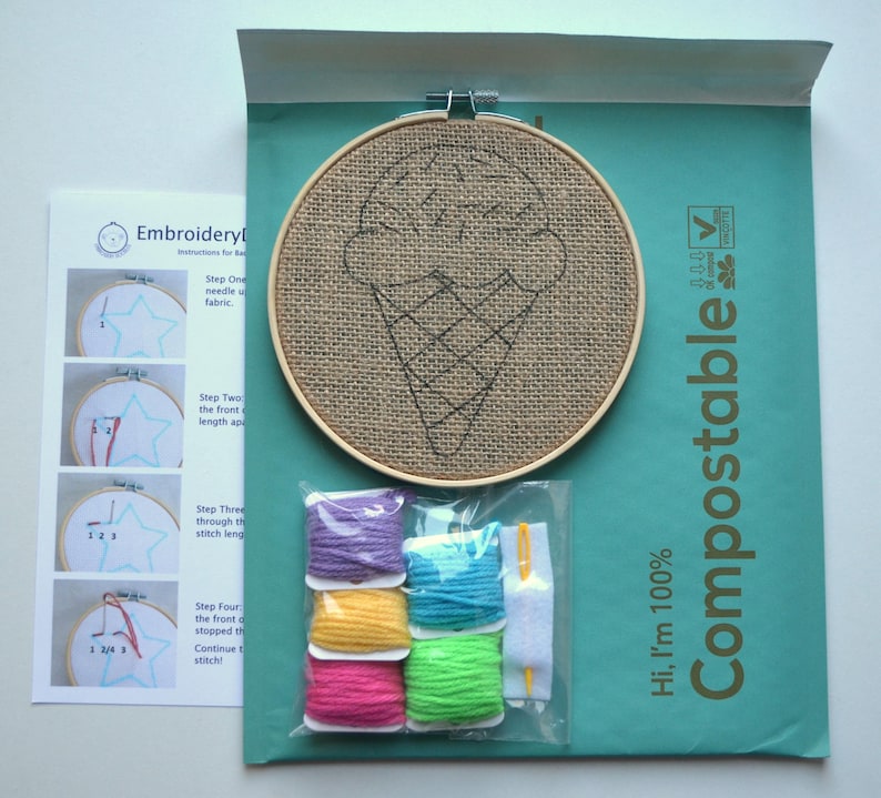 May include: A DIY embroidery kit with a pre-printed burlap hoop featuring an ice cream cone design. The kit includes four skeins of embroidery floss in purple, blue, yellow, and green, a needle, and instructions. The packaging is labeled "Hi, I'm 100% Compostable".