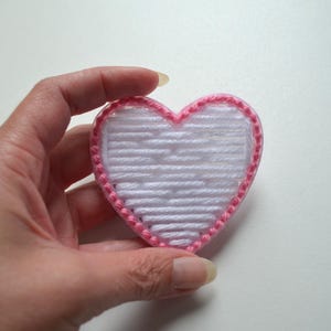 May include: A white heart-shaped yarn craft with a pink yarn border.