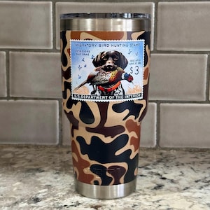 May include: Stainless steel tumbler with a camouflage pattern in shades of brown, black, and tan. The tumbler features a "Migratory Bird Hunting Stamp" design with a dog holding a pheasant. Text includes "U.S. DEPARTMENT OF THE INTERIOR".