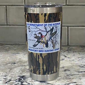 May include: A stainless steel tumbler with a camouflage pattern and a vintage U.S. Department of the Interior Migratory Bird Hunting Stamp featuring a wood duck. The stamp reads "$5 Wood Duck Void After June 30, 1975 Migratory Bird Hunting Stamp".