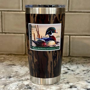 May include: A stainless steel tumbler with a camouflage pattern and a wood duck stamp featuring the text "U.S. Department of the Interior", "$15", "Wood Duck", and "Migratory Bird Hunting and Conservation Stamp".