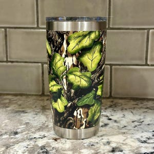 May include: A stainless steel tumbler with a camouflage pattern featuring green leaves and brown branches. The tumbler has a lid and a double-walled design.