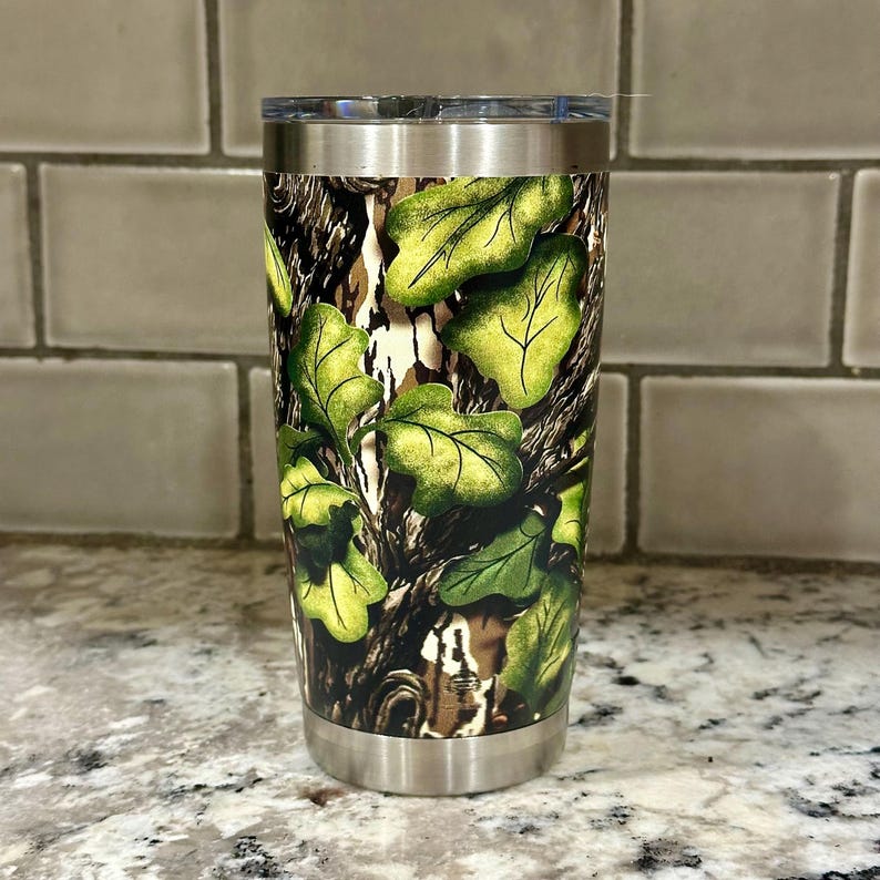 May include: A stainless steel tumbler with a camouflage pattern featuring green leaves and brown branches. The tumbler has a lid and a double-walled design.