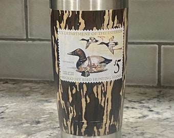 May include: A stainless steel tumbler with a brown and tan camouflage pattern. The tumbler features a vintage US Department of the Interior Migratory Bird Hunting Stamp with a canvasback duck illustration. The stamp reads "VOID AFTER JUNE 30, 1979" and "$5".