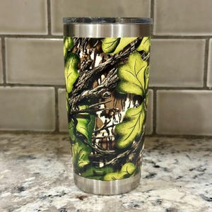 May include: A stainless steel tumbler with a camouflage pattern featuring green and brown leaves. The tumbler has a lid and is perfect for keeping drinks cold or hot.