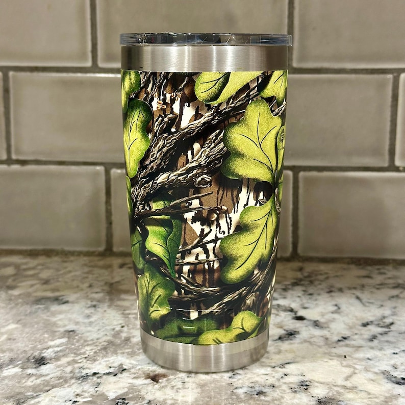 May include: A stainless steel tumbler with a camouflage pattern featuring green and brown leaves. The tumbler has a lid and is perfect for keeping drinks cold or hot.
