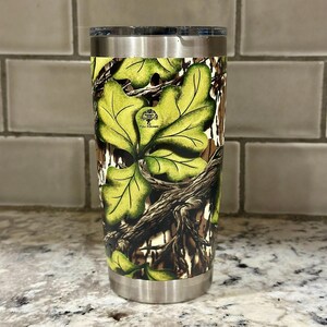 May include: A stainless steel tumbler with a camouflage pattern featuring green and brown leaves. The tumbler has a black lid and a white logo that says "Full  Pounce".
