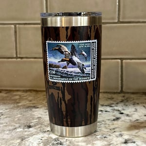 May include: A brown and black camouflage tumbler with a vintage duck hunting stamp featuring a pair of canvasback ducks. The stamp reads "Migratory Bird Hunting and Conservation Stamp" and "Void After June 30, 1983".
