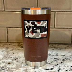 May include: A brown insulated tumbler with a stainless steel base and rim. The tumbler features a camouflage patch with the word "YETI" in black letters. The lid is clear with an orange accent.