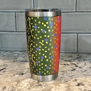May include: A stainless steel tumbler with a green, red, and yellow trout pattern. The tumbler has a lid and a handle.