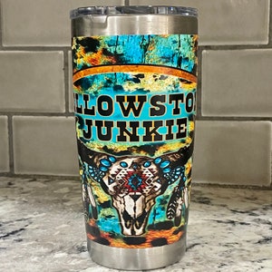May include: A stainless steel tumbler with a blue, orange, and white design. The tumbler features a skull with feathers and the text "Yellowstone Junkie".