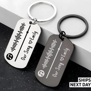 May include: Two metal keyrings with a dog tag design. One is silver, the other is dark grey. Each tag features a music waveform, a musical note icon, and the text "Our Song Emily" in a script font. The keyrings are silver and black.