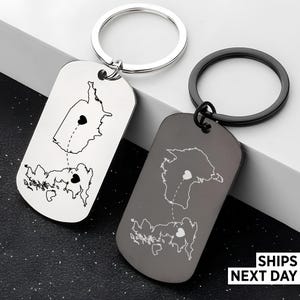 May include: Two rectangular keyrings featuring map outlines and heart symbols linked by dashed lines. One keyring is silver with a silver ring, the other is grey with a black ring. The text "SHIPS NEXT DAY" is visible in the bottom right corner.