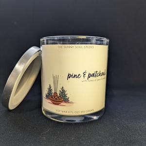May include: A clear glass candle jar with a silver lid, featuring a label with the text "pine & patchouli" and "THE SUNNY SOUL STUDIO". The label includes an illustration of pine trees and a bowl. The candle contains 9 fl oz of soy wax.