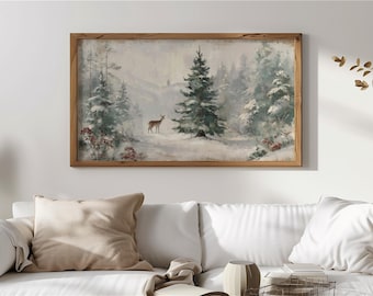 May include: A framed landscape painting depicts a snowy forest scene with a deer. The artwork features a brown wooden frame and a muted color palette of whites, greens, and browns. The painting is displayed above a white sofa with beige pillows.