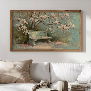 May include: A framed painting shows a park scene with a bench under a flowering tree. The artwork features a light blue bench, pink blossoms, and a soft, muted colour palette. The frame is made of wood.