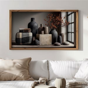May include: A framed artwork depicting a still life arrangement. It features dark grey vases, a square stone block, and a patterned cushion. Dried orange branches and small stones add texture. The artwork is set against a neutral background.