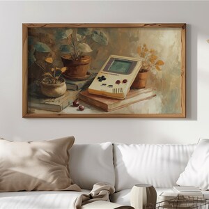 Op de afbeelding: Een ingelijst schilderij toont een vintage handheld gameconsole, potplanten en boeken. De console is crèmekleurig met rode en zwarte knoppen. Het kunstwerk heeft een warm, neutraal kleurenpalet, met een houten lijst.