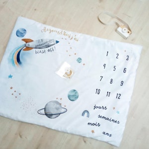 May include: A white baby milestone blanket with a rocket ship, planets, and stars. The blanket has numbers 1-12, and the words "Aujourd'hui j'ai", "blast off!", "jours", "semaines", "mois", and "ans".