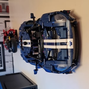 May include: A dark blue Lego sports car model with white racing stripes is mounted on a wall. The model has clear windshields and is displayed upside down. A red Lego motorcycle is visible in the background.