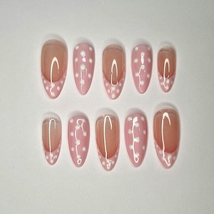 Könnte beinhalten: Ein Set aus zehn ovalen Kunstnägeln. Jeder Nagel hat eine rosa Basis mit weißen Tupfen und eine transparente, hautfarbene Spitze. Die Nägel sind in zwei Reihen auf einer weißen Oberfläche angeordnet.