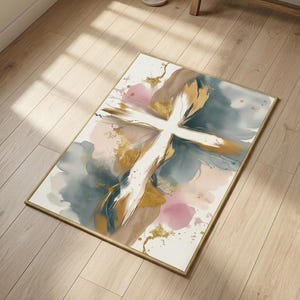 Puede incluir: Una alfombra rectangular con un diseño abstracto que presenta una forma de cruz blanca central. El patrón incluye lavados de acuarela mezclados en tonos azul polvoriento, rosa suave y bronceado, acentuados con salpicaduras de pintura dorada.