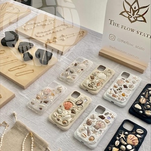 Puede incluir: Una colección de fundas para teléfonos decoradas con conchas marinas. Las fundas son de varios colores, incluyendo blanco, negro y beige. Las fundas están dispuestas sobre una superficie blanca. El texto "THE FLOW STATE" es visible en un cartel en el fondo.